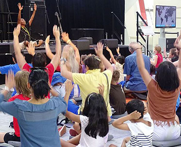 Woman sitting on a stage with audience of men, women and children sitting on the floor, raising their hands above their heads.