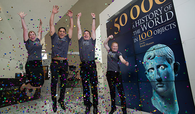Two women and two men in uniform jumping in the air amidst confetti, adjacent to a promotional billboard.