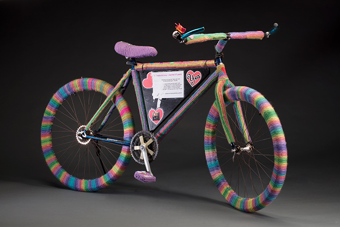 A bicycle covered with woollen crochet that consist of multi-coloured striped wheels, a purple seat and two purple peddles.