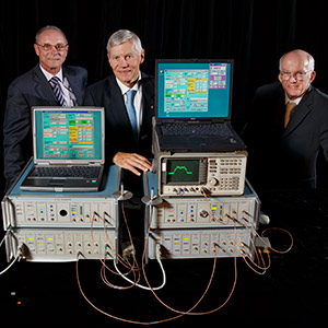 three men in suits standing beside a table on which sit a collection of cumbersome electronic equipment.