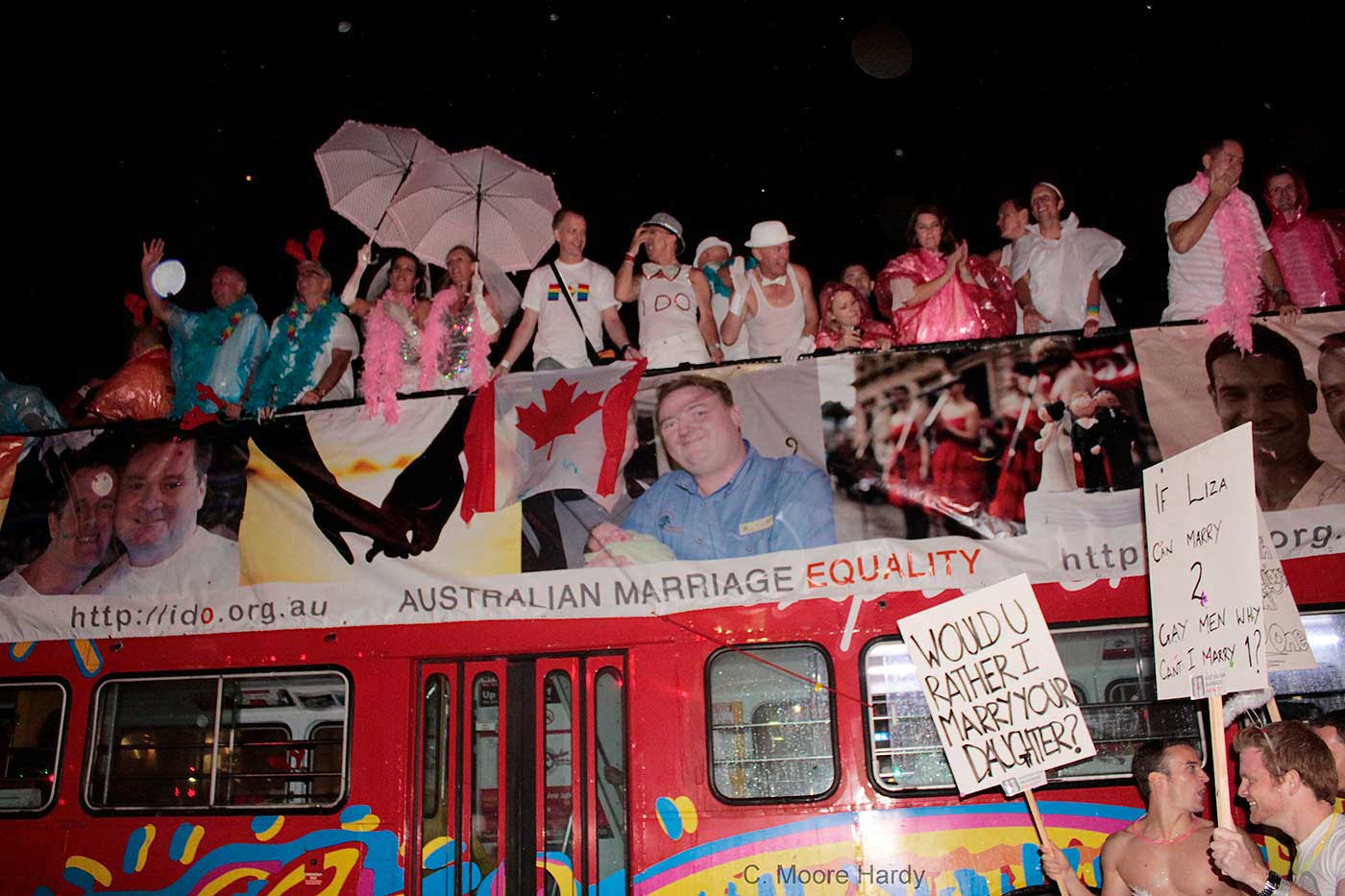 Red open-top double-decker bus with crowds of people in pink and other colours on the top deck.