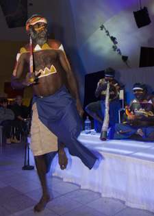 A colour photograph of an Indigenous man engaged in a traditional dance. He wears a sarong-style garment around his lower body and has traditional patterned markings on his upper body. Behind him on a low stage sit two other Indigenous Australian men. One playing the didgeridoo while the other plays a pair of clapping sticks. Both have traditional markings on their upper bodies.