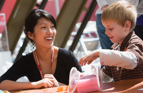 A colour photograph of a female museum staff member and a young boy sitting at a table. He has a toy car on the table, made from craft materials.