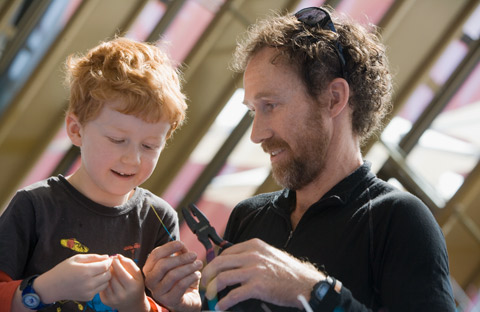 A father and his young son engaged in a craft activity. The father holds a pair of pliers in his left hand. His son is to the left and appears to be holding something small between the thumb and forefinger of each hand. Behind them, out of focus, the background has a pattern of bronze-coloured diagonal stripes.