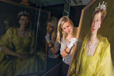 Curator Laura Breen, wearing white gloves, peers around a framed portrait of Queen Elizabeth II. The scene is reflected in a window at the left of the image.