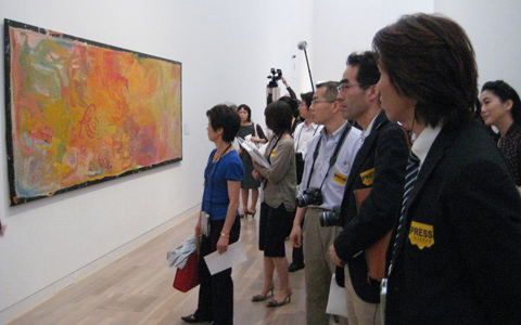 A painting in pastel colours of yellows, pinks, blues and greens displayed in landscape orientation is on the left side of the image; nine Japanese viewers range along the right side of the image. One of the viewers in the background of the image holds a camera above their head.