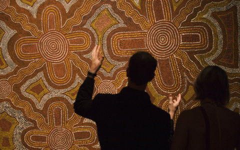 This image is shot from behind with two people viewing an Aboriginal dot painting in ochre colours. All of the light in the image is on the painting; both people are in silhouette with only their outlines visible, except that the man who is on the left is gesturing towards the painting and his hands are in the light.