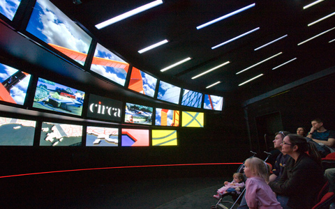 Seven people sit in a darkened room in the front seats of a theatre. There are 18 screens ranged in three rows with a variety of images and a series of slanted light areas on the ceiling. On the centre screen is the word 'circa' in white text on a black background.