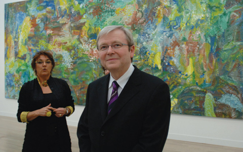 Kevin Rudd is at the centre-front of the image facing the camera. To the left of the image and further back stands curator Margo Neale. In the background is a large painting in greens and blues with yellow highlights.