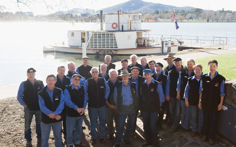 Group of 23 people standing in front of the paddle steamer at its dock. The lake and hills can be seen in the background.