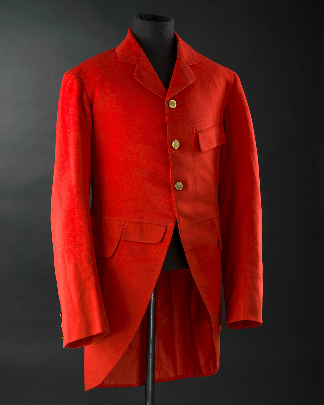 A red wool cut away jacket with tails otherwise known as hunting pinks. It has 3 brass shank buttons at front opening and 3 buttons on each cuff. It is fully lined in red cotton. The sleeves are lined in white cotton with blue stripe. The jacket has machine quilted underarm lining and a back vent with 2 brass buttons at top. It has four pockets on front with flaps. - click to view larger image