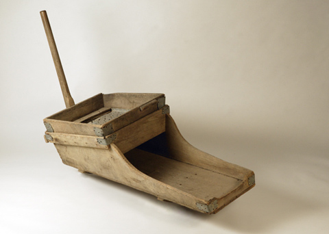 A gold washing cradle made of wood with metal fixtures. The main body of the cradle is supported by two curved pieces of wood, one at either end, with bolts in them that then fit into holes in a rectangular wooden frame that the cradle sits on top of. The body of the cradle is higher at one end, to accommodate the sieve and the tray that sits below the sieve to catch the gold. Both of these components are roughly square in shape. The sieve is made from a sheet of metal with holes punched in it.