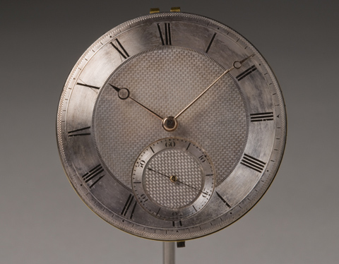A chronometer with a silver-coloured face featuring two hands and Roman numerals around the edge. A smaller dial with two hands and numbers 10 - 60 in tens is at the bottom of the face.