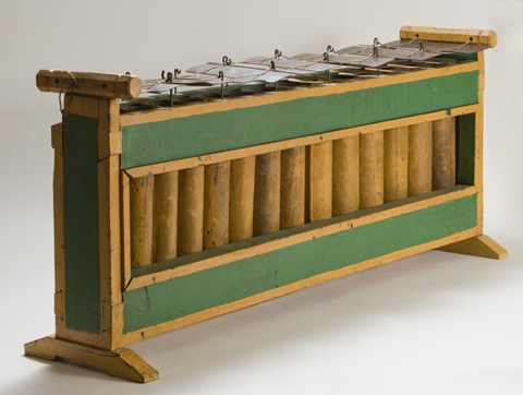 A wooden instrument featuring fourteen metal cylinders, painted yellow and encased in a rectangular wooden frame. There are fourteen rectangular metal slats on the top, of varying sizes and strung together with string, which is suspended from metal hooks. The wooden case is painted yellow and green.