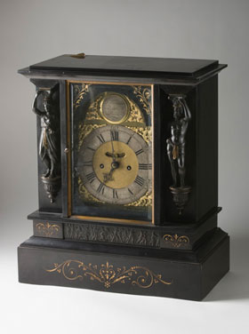A mantelpiece clock, showing the face and one of the sides. The clock is made from dark material and has carved decorations.