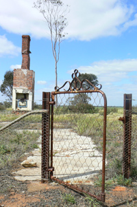 Rusted metal gate at the front of old house foundations. A brick chimney appears at the rear of the image.