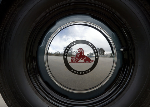 Close up shot of a shiny silver hubcab bearing the lion and 'General Motors Holden' logo. The hubcab is framed by tyre rubber and reflects a cloudy blue sky and gum trees.