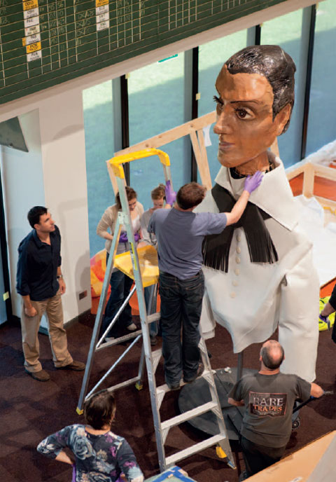 Photograph taken from above, looking down at an oversized puppet consisting of a female face, constructed of painted papier-mache. The woman wears a white collared-shirt and black taselled scarf. A man on the third rung of a large ladder reaches up to adjust the puppet's collar. Five other people stand in the gallery space.