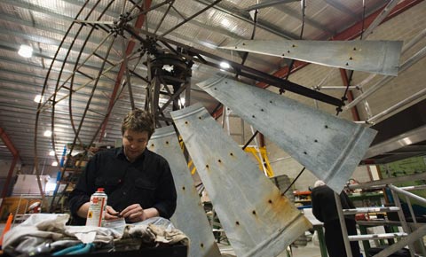A man stands at a workbench, holding a small piece of metal. Behind him towers a partially re-assembled windmill.