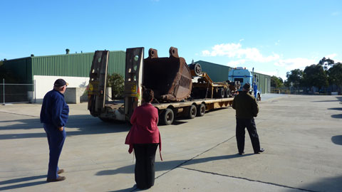 A view of a large open concreted area in front of two large green industrial sheds. In the foreground, three people (two men and a woman) stand with their backs to the camera, looking at a semi-trailer in the centre of the image. The rear of the semi-trailer is closest to the people. The semi-trailer prime mover is visible at the far end of the trailer. On the tray of the semi-trailer is the large metal bucket from an excavator used in an iron ore mine. The bucket sits facing the rear of the trailer. There is some other mining equipment on the trailer, between the bucket and the prime mover. The bucket is a dark earth colour, stained by its use in the mine. Above the two large industrial sheds is blue sky and some low clouds visible in the right background.