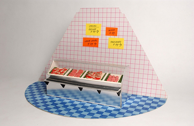 A diorama of the interior of a Butcher's shop, consisting of a blue checkered patterned semi circle cardboard base, a pink and white checkered triangular carboard background and a cardboard and plastic display case filled with modelling compound 'steaks' and 'sausages'.