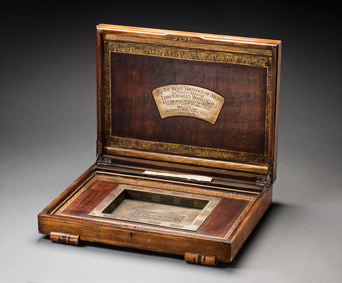 A wooden box with a raised lid. An engraved metal plaque attached to the underside of the lid, conveys the details: ‘TO THE MOST VIRTUOUS OF MEN / VIA “FAITH IN AUSTRALIA” / LORD CHARLES WAKEFIELD / SYDNEY-NEW PLYMOUTH TRANS / TASMAN FLIGHT 7TH DEC 1933 / PAR AVION FROM / ROSLYN FOSTER BOWIE PHILP / QUEENSLAND’. The inscribed date reflects Ulm’s changing departure dates.
