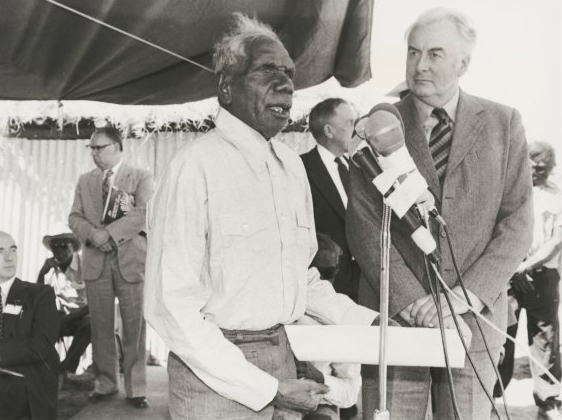 Aboriginal man holding document speaks in front of microphones on stand, with Prime Minister Whitlam to his left.