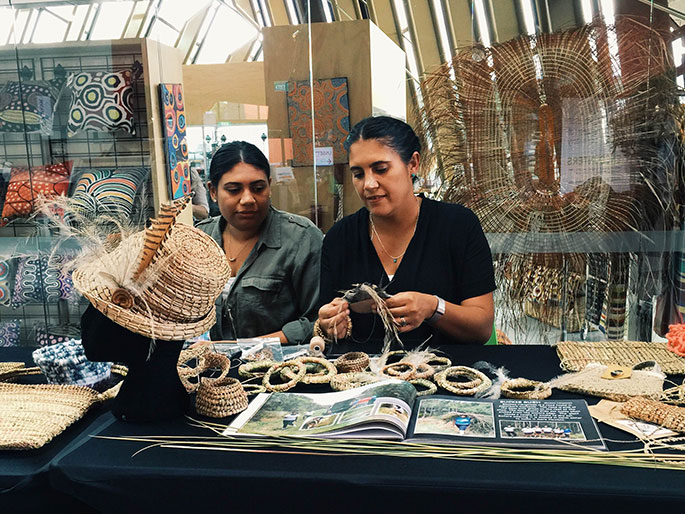 Two women sitting at a table with various woven objects