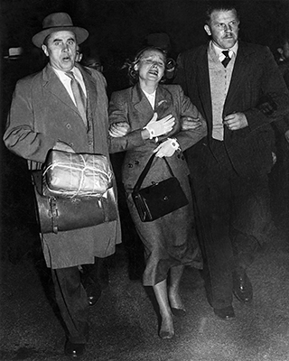 Photo taken at night showing distressed woman flanked by two men in suits holding her arms