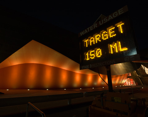 An electronic sign outside a museum, at night. The sign is at the right of the photograph. It has a display which is headed 'Water Usage', in white letters. Under the heading, in a light readout, is 'Target 150 ML'. The letters in the readout are yellow. Behind the sign is part of the museum's exterior. It is a curved wall that is lit up by a row of lights in the ground. The wall is orange in colour. To the far left of the photograph can just be made out what appears to be the entrance to the museum.
