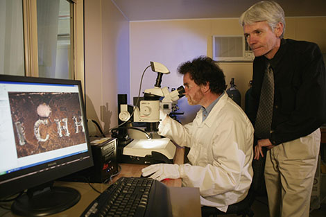 One man wearing a white coat and gloves sits side-on in the centre of this image, looking into a telescope. To his left a computer screen displays a section of brass plate, stamped with the letters 'ICHH'. A second man stands behind the first, viewing the computer screen.