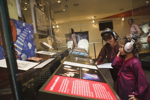 Two young girls wearing headphones stand beside a case containing a painted board and other objects.