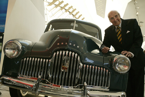 Jack Rawnsley leans on the front righthand side bonnet of a dark blue Holden. The silver grille and headlamps are prominent. The Museum's Hall forms a backdrop.