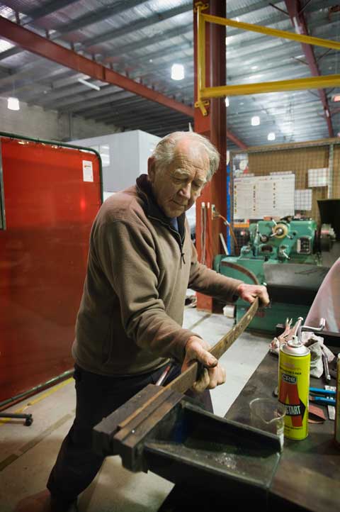 A man moves his weight towards a long, narrow strip of metal which is held in place at one end by a vice.