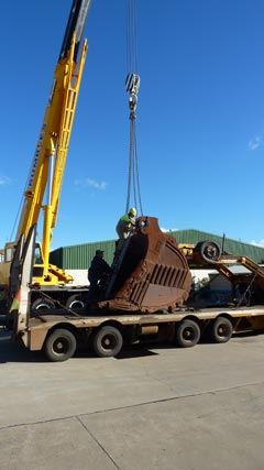 A view of a large mining excavator bucket being prepared for lifting off a semi-trailer. At the left-hand side of the image is a large yellow crane, facing the semi-trailer. The bucket faces the rear of the semi-trailer, which is at the left-hand side of the image. A man has climbed up a ladder positioned at the front of the bucket and is attaching chains from the crane. A second man is at the base of the ladder, steadying it for the other man.