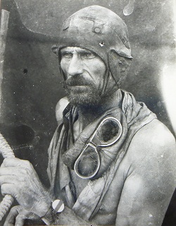 Black and white image showing a bearded and bare-chested man gazing to the left of frame. He wears a leather flying-style cap and has goggles draped around his neck. His right arm clutches a rope.