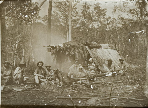 Black and white glass plate negative showing eight men sitting in front of a shelter in the bush. The A-frame shelter is made of bark and leaves and has a billy can hanging at the front. The men are gathered around a smoking fire, with another billy can hanging over the it. A dog sits at the front and centre of the image.