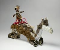 A white, grey and brown model camel with two riders made from, raffia, minarri (greybeard) grass, wool and leather. The two riders are seated on a red crocheted saddle, which is fastened to the camel with leather straps and leads. 