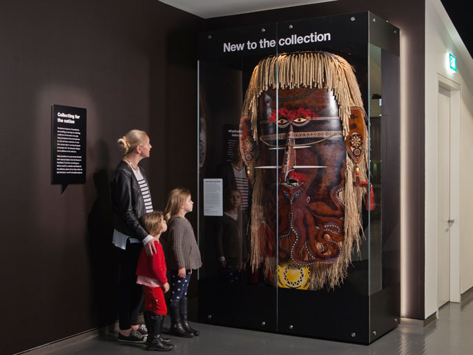 A woman and two small girls stand to the left, looking at a large mask on display in a perspex case under the heading 'New to the collection'.