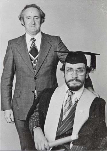 Black and white photo showing a seated man wearing academic gown and hat. A man in s suit stands to his left.