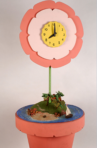 A large clock in the shape of a flower in a pot. The pot and petals are painted pink and the stem is painted green. A large yellow clock face is positioned in the centre of the flower head, which has black plastic numbers and hands.