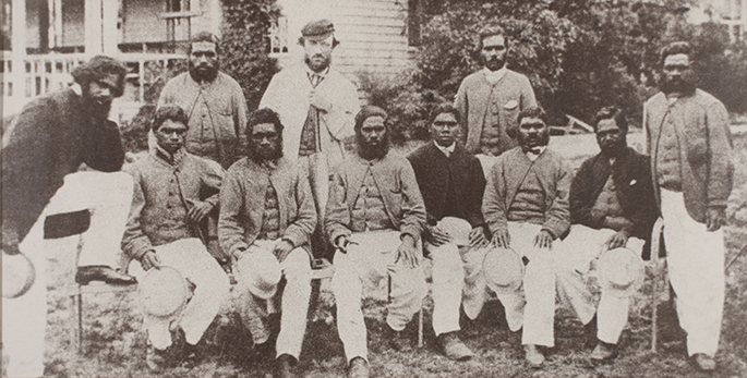 Black and white reproduction photograph of a cricket team of eleven men, six of whom are seated. They are outside in front of a building.