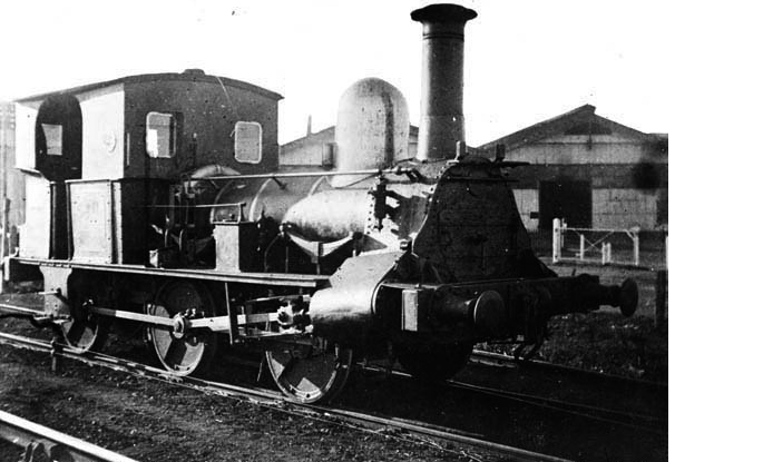 Slightly scratched photo of an early steam engine with railway sheds in the background.