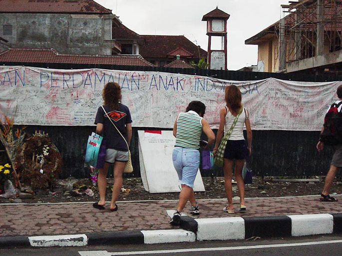 Three young women with their backs to the camera look at a memorial consisting of a long white sheet covered in handwriting, pinned to a fence above some wreaths and flowers.