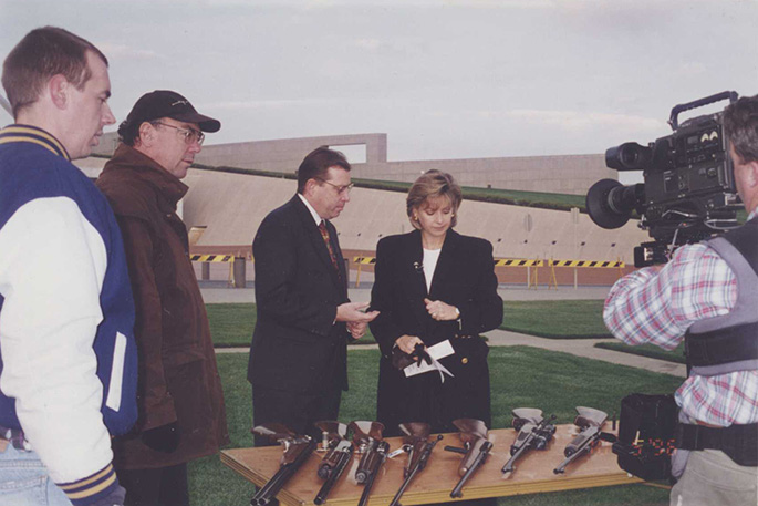 A person is filming a group of people looking at several guns on top of a table outside Australian Parliament House.