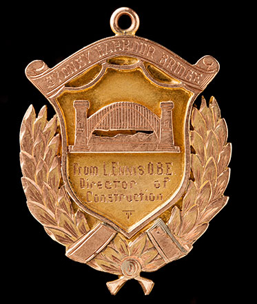 Faded gold-coloured metal badge with an image of the bridge and the words ‘From L Ennis OBE, Director of Construction’.