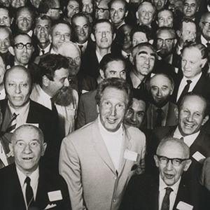 interior photo of tightly packed group of about 70 men in their 50s and 60s smiling at the camera.