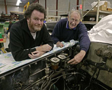 Two men lean on the side of a car, with its bonnet removed and engine bay exposed.