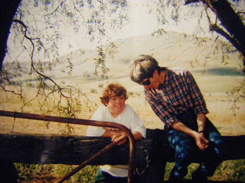 A woman on the right sits on a timber post fence, leaning down towards a young girl, who stands, resting her arms on the fence. The image is framed by tree leaves, with a metal gate in the left foreground and a small hill rising in the background.