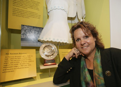 Evonne Goolagong Cawley sits beside a case containing a small circular trophy, white tennis dress, text panels and black and white photograph.